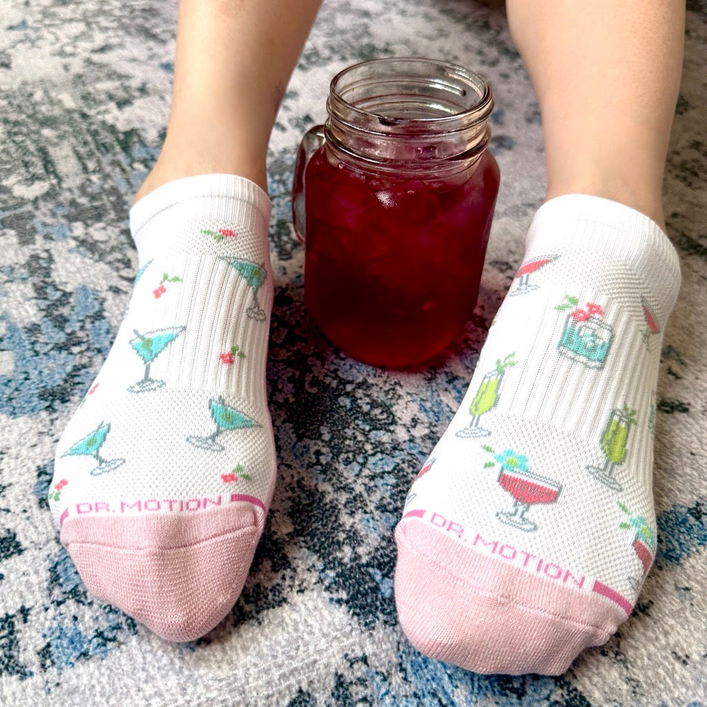 White socks with cocktail pattern and pink toes worn by a person, sitting on a textured surface next to a jar of red liquid.