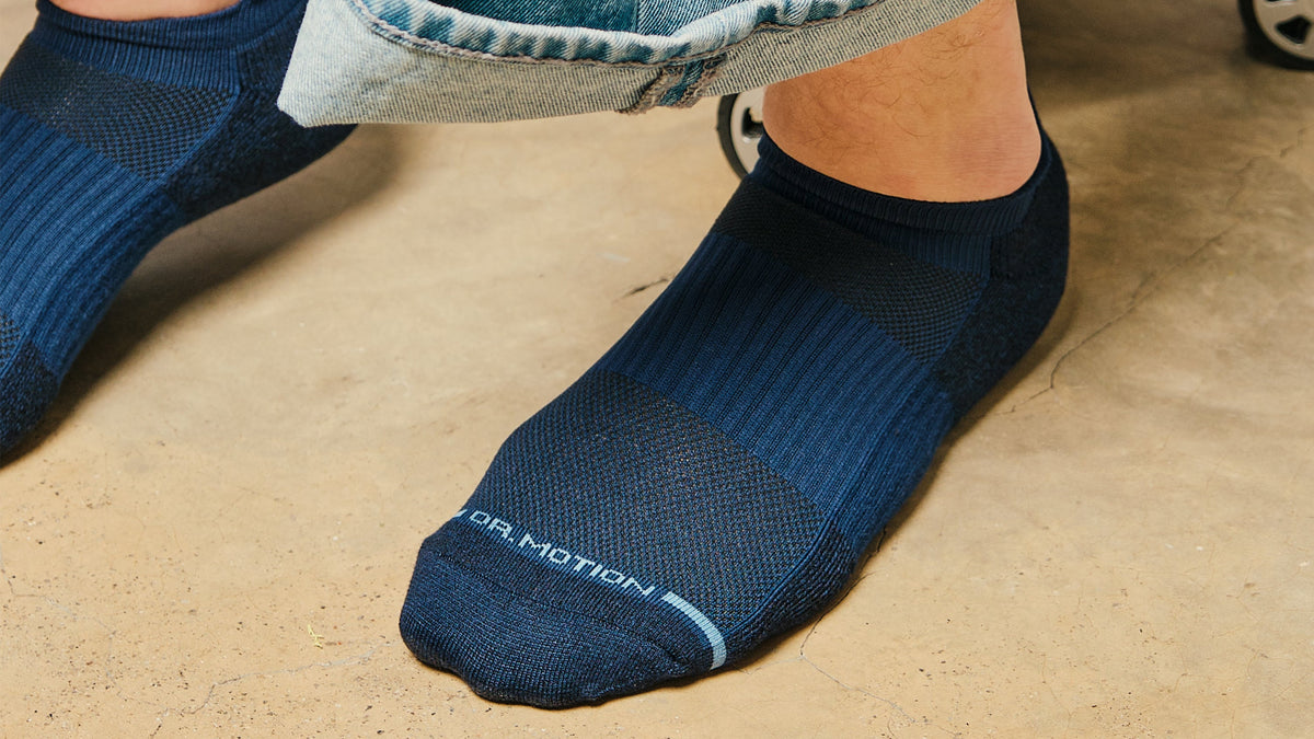 Navy ankle compression socks worn with rolled-up denim pants, next to a silver suitcase on a concrete surface.