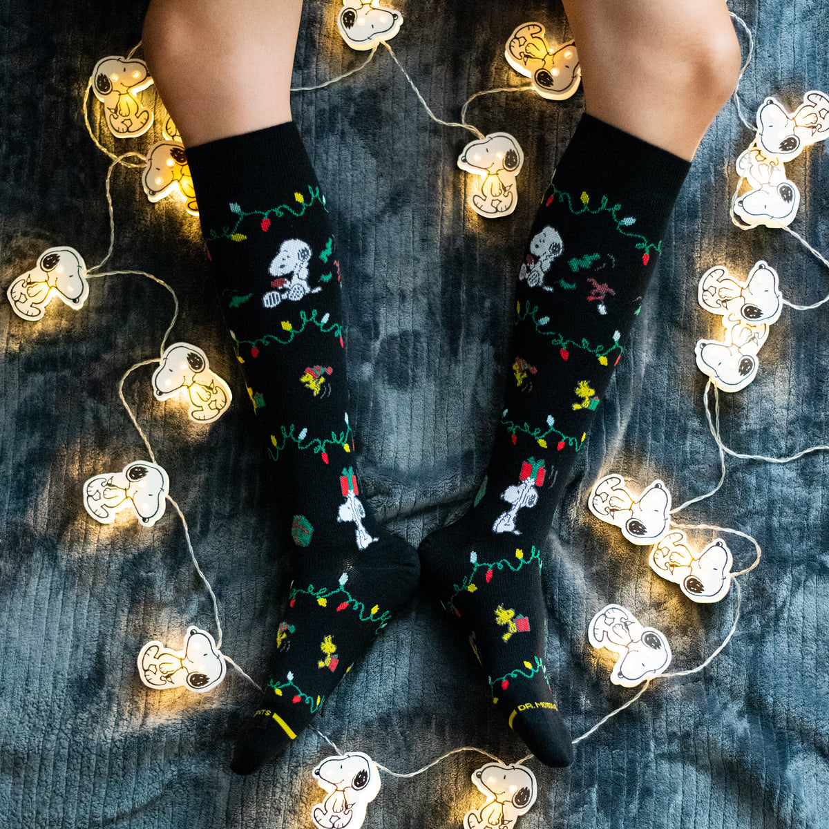 Black knee-high compression socks with Snoopy and holiday light designs, worn on a cozy blanket with string lights.