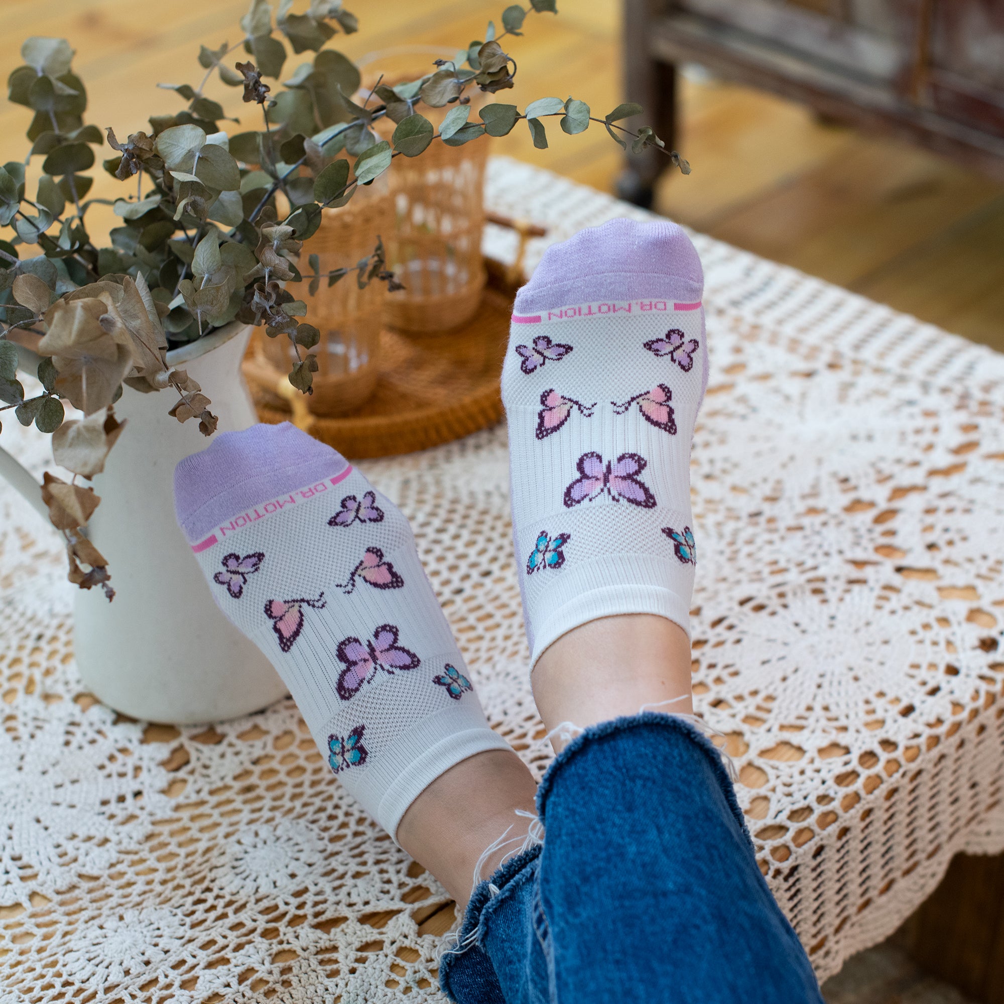 Ankle compression socks with butterfly designs in pink and blue, worn with denim jeans on a lace tablecloth.