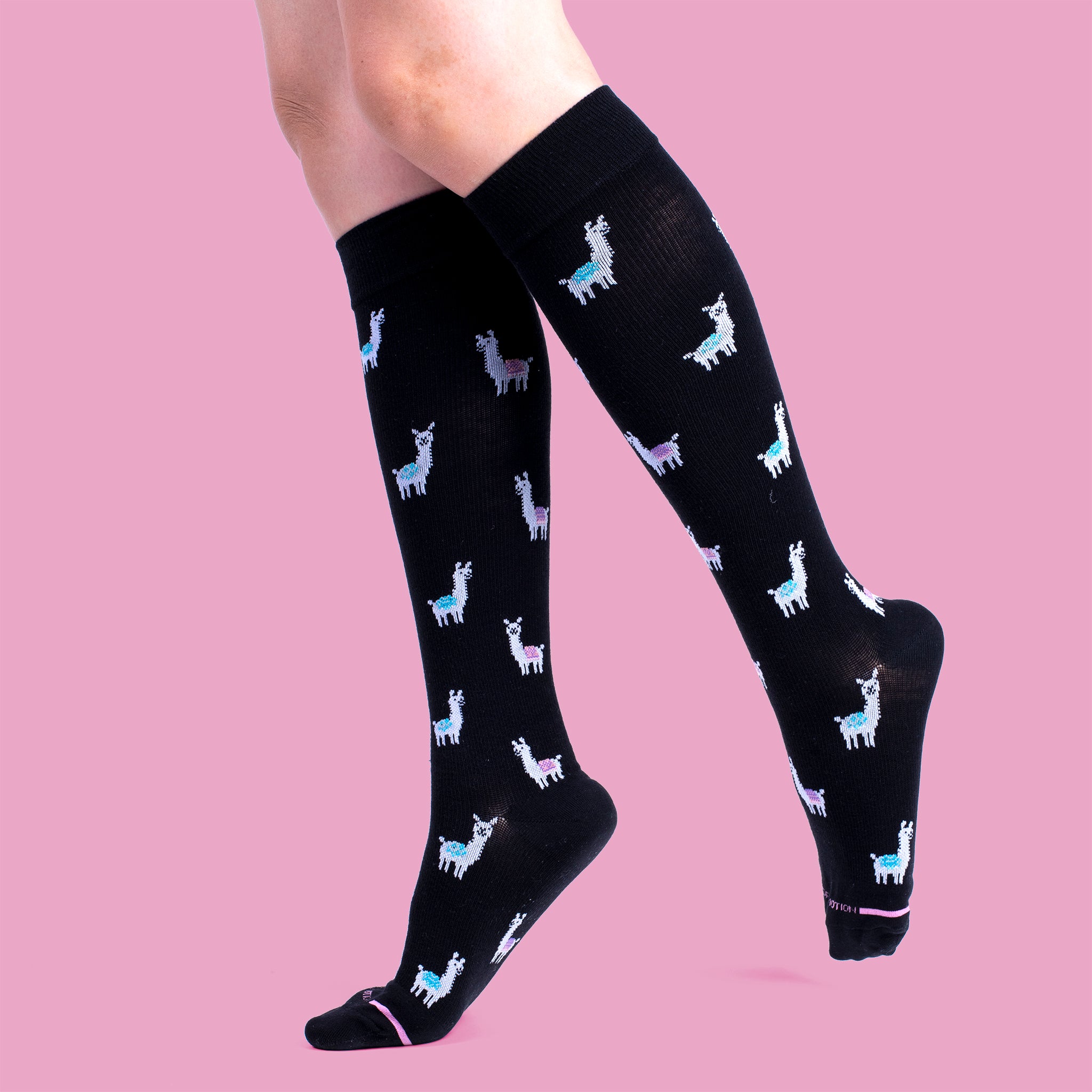 Knee-high black compression socks with colorful llama patterns, worn on a model against a pink background.