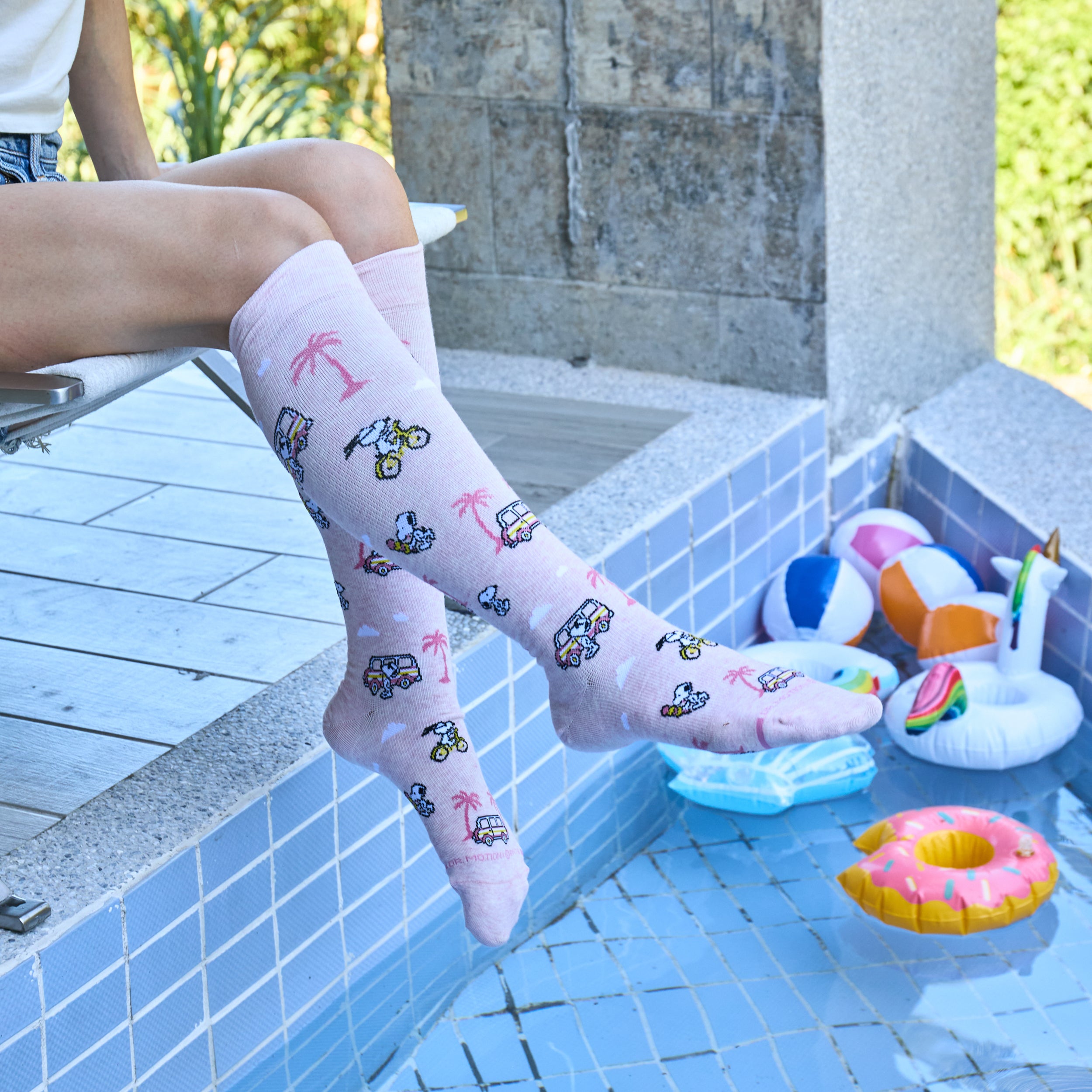 Knee-high pink compression socks with Snoopy and beach-themed designs, worn by a person sitting by a pool.